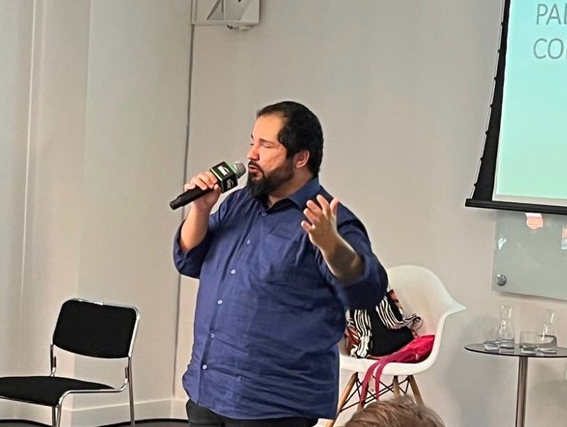 homem pardo com cabelos e barba pretos, vestinfo uma blusa social azul e calça jeans da uma palestra segurando microfone